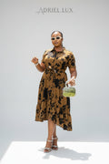 Woman in a patterned dress holding a green handbag against a white wall with 'Adriel Lux' branding.
